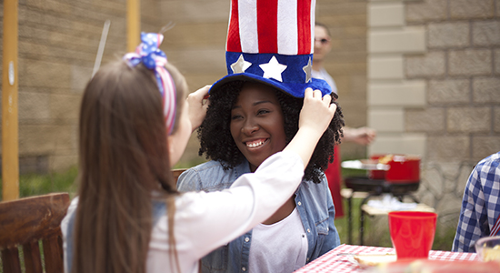 A young girl puts a patriotic novelty hat onto a young woman's head - outside at a picnic